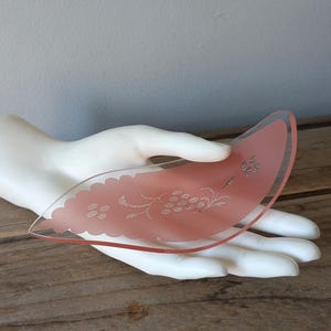 May include: A pink, fan-shaped glass dish with a floral etched design. The dish is held by a white hand sculpture, set against a rustic wooden surface. The glass has a delicate, translucent quality.