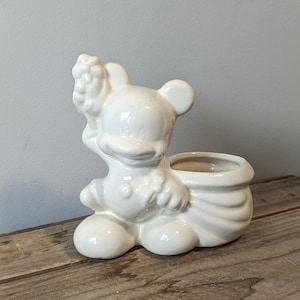 May include: White ceramic planter shaped like a cartoon character holding a flower. The planter has a large pot on the back for planting.