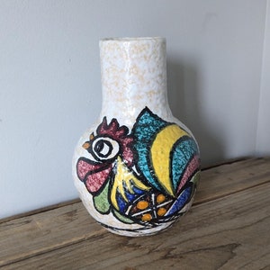 Puede incluir: Un jarrón de cerámica blanco con un diseño de gallo. El gallo está pintado en tonos de rojo, amarillo, azul y verde. El jarrón tiene una superficie texturizada.