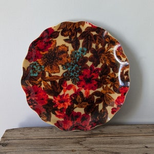 May include: A decorative plate with a floral pattern. The plate has a beige background with red, orange, teal, and brown flowers and leaves. The plate has a scalloped edge and is on a wooden crate.