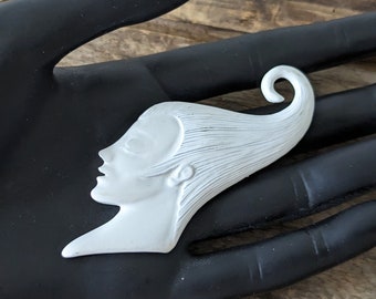 Puede incluir: Un broche de cerámica blanca con forma de perfil de mujer con el pelo suelto. El pelo está peinado en un recogido caprichoso con un rizo en el extremo.