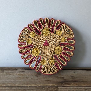 Könnte beinhalten: Ein runder geflochtener Untersetzer mit rotem und gelbem Design. Der Untersetzer ist aus natürlichen Materialien gefertigt und hat ein geometrisches Muster.