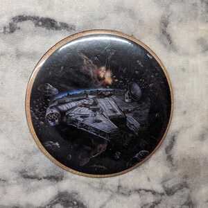 May include: A round, black and white button featuring a spaceship in space. The spaceship is flying through a field of asteroids with an explosion in the background.