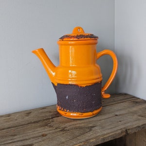 May include: Orange and brown ceramic teapot with a textured finish. The teapot has a rounded body and a spout on the side. The lid is slightly domed and has a small knob on top.