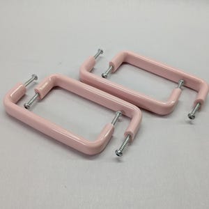 Puede incluir: Dos tiradores de armario rectangulares con un acabado en rosa pastel suave. Cada tirador presenta un diseño de marco redondeado y liso con dos tornillos metálicos plateados unidos a los puntos de montaje, listos para su instalación.