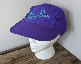 May include: Purple nylon baseball cap with a teal embroidered Ray-Ban logo.
