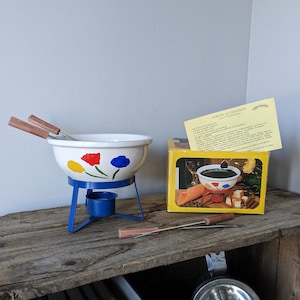 May include: A white ceramic fondue pot with a blue metal stand and wooden handles. The pot has a floral design with red, yellow, and blue flowers. The pot is in its original box, which is yellow with black text. The box includes instructions for making chocolate fondue.