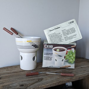 May include: A white ceramic fondue pot with a black and yellow striped design. The pot is on a white stand with holes for heat. Four wooden handled fondue forks are included. The box says "OBO Chocolate Fondue Set Ensemble a Fondue au Chocolat".