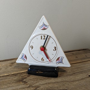 May include: White ceramic triangular clock with gold numbers and hands. The clock has a black base and features a decorative design on each side.