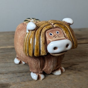 May include: A ceramic figurine of a brown Highland cow with a white face and white hooves. The cow has a brown and yellow mane and a small green flower on its head.