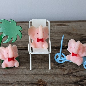 Op de afbeelding: Drie roze pluche olifantjes met rode strikjes. Eén olifantje zit op een witte stoel, één zit op een blauwe scooter en één staat onder een groene palmboom.