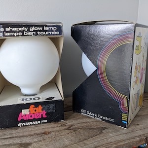 May include: Two boxes for a 100 watt light bulb. The box on the left is black with white text that reads "The shapely glow lamp Une lampe bien tournée 100 fat Albert SYLVANIA". The box on the right is black with rainbow stripes and white text that reads "GTE Sylvania Canada Drummondville, Quebec".