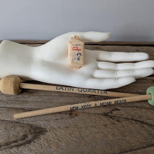 Op de afbeelding: Een witte keramische hand houdt een klein, rechthoekig pakketje met tekst en een cartoonafbeelding vast. Er zijn ook twee houten drumstokken met de tekst "Latin Quarter" en "New York & Miami Beach". Eén drumstok heeft een groene kop.