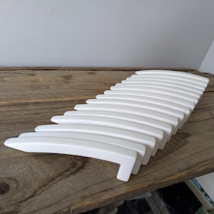 May include: A white, curved decorative object with a series of parallel, arched elements. The object is made of a smooth, glossy material and is arranged on a rustic, wooden surface. The design is modern and minimalist.