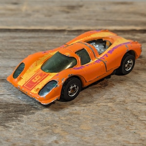 May include: Orange and yellow vintage toy race car with a green windshield. The toy car has black wheels and a silver engine. The car has a red and yellow stripe on the front with the number 3.