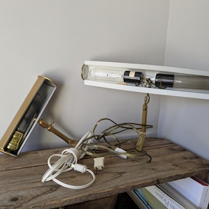 May include: A vintage desk lamp with a white rectangular shade and brass accents. The lamp features a long, cylindrical light bulb and a separate, matching gold-colored base. The lamp is on a wooden surface.