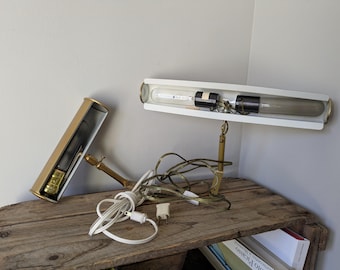May include: A vintage desk lamp with a white rectangular shade and brass accents. The lamp features a long, cylindrical light bulb and a separate, matching gold-colored base. The lamp is on a wooden surface.