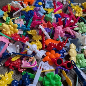 Puede incluir: Una colección de coloridos accesorios para el cabello de plástico, que incluyen horquillas, pinzas y pasadores. El surtido presenta varias formas como flores, lazos y figuras de dibujos animados en tonos rosa, amarillo, azul y naranja. Algunos clips tienen elementos decorativos.