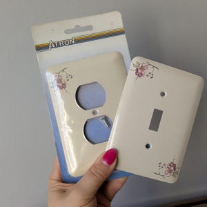 Op de afbeelding: Off-white stopcontact- en lichtschakelaar covers met bloemaccenten. De stopcontact cover heeft twee gaten voor stekkers en de schakelaar cover heeft een rechthoekige opening. De covers zijn nieuw en in de originele verpakking.