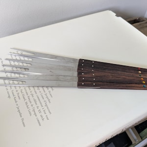 Può includere: Un set di sei coltelli da formaggio in acciaio inossidabile con manici in legno scuro. Ogni manico è adornato da un piccolo punto colorato. I coltelli sono disposti su una pagina di testo, probabilmente di un libro.