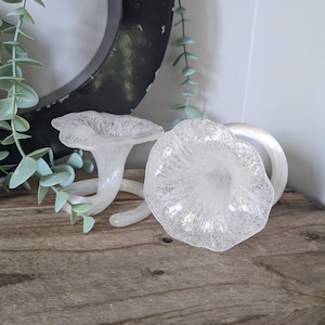 May include: Two white glass trumpet-shaped vases with a textured surface. The vases are on a wooden surface.