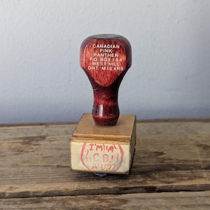 May include: A vintage wooden stamp with a dark red handle and a rectangular base. The stamp reads "CANADIAN PINK PANTHER" and other text. The base has a faded red stamp that reads "I'M A C.B. NUT".