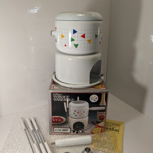 Op de afbeelding: Een witte Colibri 11-delige fondue-set. De fonduepot heeft een deksel en is versierd met kleurrijke driehoeken. De set bevat een standaard, vorken en andere accessoires, allemaal in een doos.