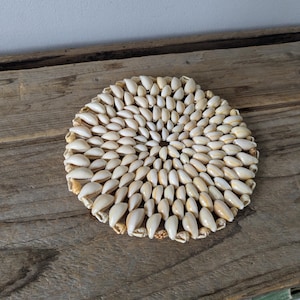 Może przedstawiać: Okrągły podkładka pod szklankę wykonana z małych, kremowych muszli ułożonych w okrągły wzór. Muszle są ciasno upakowane, tworząc teksturowaną powierzchnię. Podkładka leży na zwietrzałej drewnianej powierzchni, dodając rustykalnego charakteru. Przedmiot dekoracyjny do domu.