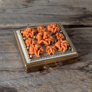 Peut inclure: Un poudrier vintage avec un cadre en métal doré et un couvercle en émail blanc. Le couvercle est décoré d'un motif floral avec des fleurs de couleur corail.