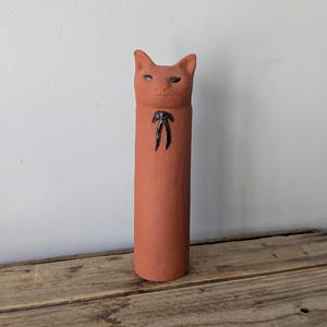 Può includere: Statuina di gatto in ceramica color terracotta. Il gatto ha un corpo cilindrico con una faccia di gatto in cima. La faccia del gatto ha piccoli occhi scuri e un papillon nero. La statuina è appoggiata su una superficie di legno.