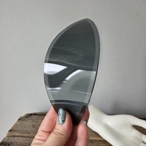 May include: A dark gray, teardrop-shaped decorative glass piece with a reflective surface. The glass has a layered appearance with lighter gray and white accents. The object is held in a hand with painted nails, and a white mannequin hand is in the background.