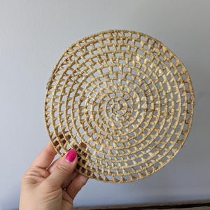 Könnte beinhalten: Ein runder, gewebter Platzdeckchen aus naturfarbenem Stroh. Der Platzdeckchen hat ein Spiralmuster mit einer Reihe konzentrischer Kreise. Das Stroh ist dicht gewebt und erzeugt eine strukturierte Oberfläche. Der Platzdeckchen hat einen Durchmesser von etwa 25 cm.