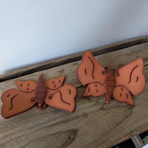 Puede incluir: Dos adornos decorativos de mariposa. Las mariposas son de color marrón rojizo con detalles marrón oscuro. Están sobre una superficie de madera envejecida, con una pared blanca al fondo.