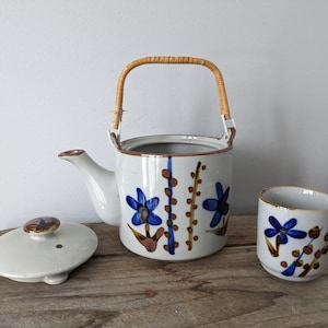 Puede incluir: Una tetera de cerámica blanca con un diseño floral marrón y azul y un asa tejida. La tetera tiene una tapa a juego y una taza pequeña con el mismo diseño.