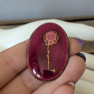 May include: Oval burgundy resin pendant with a gold flower design. The pendant has a wooden frame and a glossy finish. The flower has a detailed outline and a stem. The artist's initials are visible in the lower right corner.