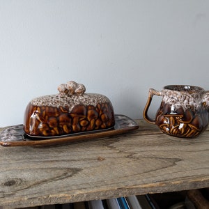Op de afbeelding: Een bruine en witte keramische botervloot en roomkan met een paddenstoelontwerp. De set staat op een houten oppervlak.