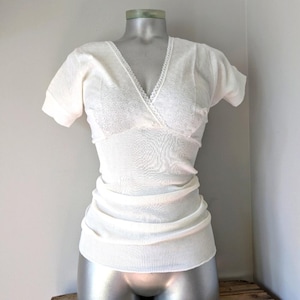 Puede incluir: Top de color crema, de manga corta, con cuello en V y silueta entallada. El top presenta un delicado ribete de encaje a lo largo del escote y una textura acanalada en la parte inferior. La prenda se exhibe en un maniquí.