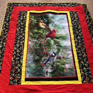 May include: A quilted throw blanket with a red, yellow, and black border. The center panel features a colorful image of three birds perched on a pine branch with red berries.