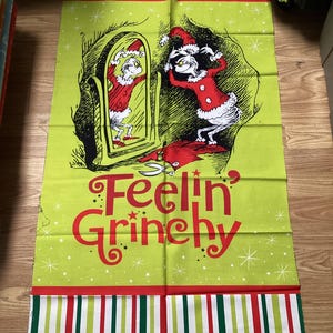 May include: A rectangular fabric panel featuring a cartoon Grinch character in a red Santa suit, looking in a mirror. The panel has a light green background with snowflakes and the words "Feelin' Grinchy" in red. The top and bottom borders have red, white, and green stripes.