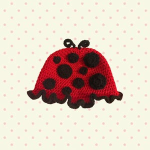 May include: A red crocheted hat featuring black circular spots of varying sizes across the surface, two small black loops at the top, and a ruffled black crocheted edge along the bottom hem, designed with a ladybug-inspired aesthetic.