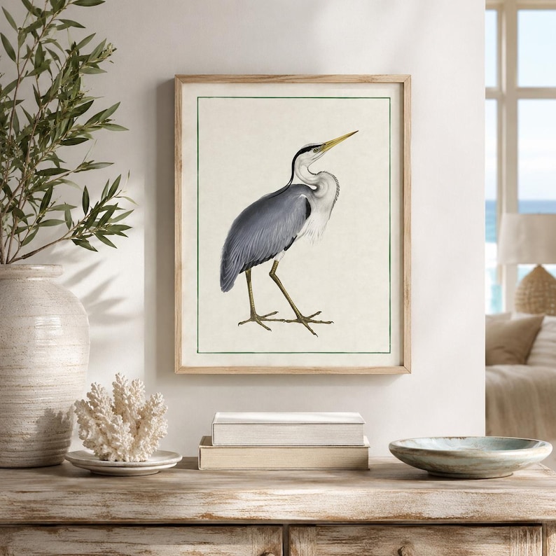 May include: A framed wall art print featuring a detailed illustration of a grey heron standing in profile. The bird has grey and white plumage, a long yellow beak, and thin legs. The artwork is set against a cream background with a thin green border.