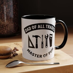 May include: A white ceramic mug with a black interior and handle. The front features black text reading DAD OF ALL TRADES and MASTER OF PUN, with black graphic icons of a triangle ruler, hammer, screwdriver, wrench, and hand saw in between.