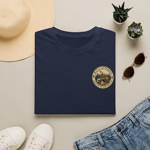 Pode incluir: Uma t-shirt azul-marinho com um gráfico circular no peito. O design retrata uma paisagem de montanha com uma cafeteira, uma caneca e o texto "WILD & CAFFEINATED" e "SURVIVING THE HOME FRONTIER" dentro de uma borda circular.
