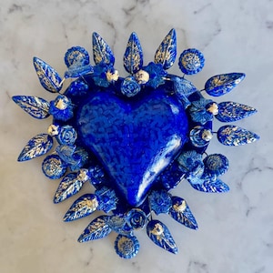 May include: A cobalt blue heart-shaped decorative object with a textured surface, surrounded by blue leaves and flowers with gold accents. The heart is the focal point, with the floral elements arranged around it. The piece is set against a marble background.