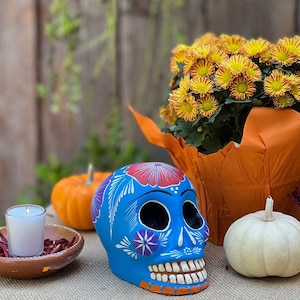 May include: A blue Day of the Dead skull with floral designs, orange pumpkins, a white pumpkin, and yellow flowers in an orange pot. A lit candle sits in a terracotta bowl with red petals.