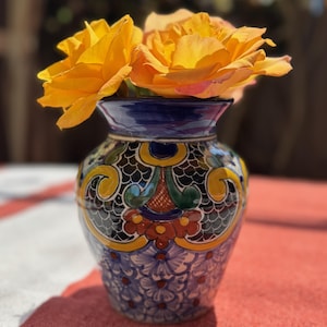 May include: A vibrant, hand-painted ceramic vase with a blue rim and intricate floral patterns in yellow, green, and red. The vase holds two large, bright yellow roses, creating a striking contrast.