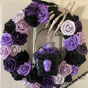 May include: A Halloween wreath featuring a circular arrangement of black, purple, and lavender roses. A black basket with a purple skull and a decorative bird is suspended in the center. A skeleton hand and a black bird are also included.