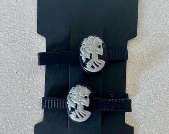 May include: Two black hair clips with oval cameos featuring a white skull design. The clips are attached to a black card with a hang hole. The cameos have a black background and a white skull profile.