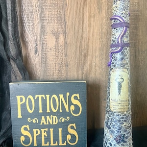 May include: A black wooden sign with gold lettering that reads "Potions and Spells".  Next to the sign is a glass bottle with a purple snake wrapped around it. The bottle has a label that reads "Snake Venom" and a description of its use.