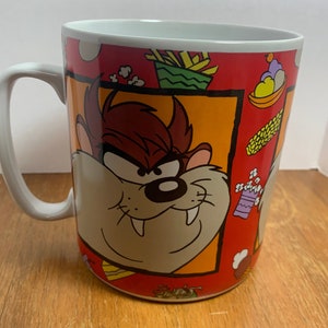 May include: A ceramic coffee mug featuring the Tasmanian Devil cartoon character. The mug has a white handle and a colorful design with food illustrations. The mug is approximately 4 inches tall.